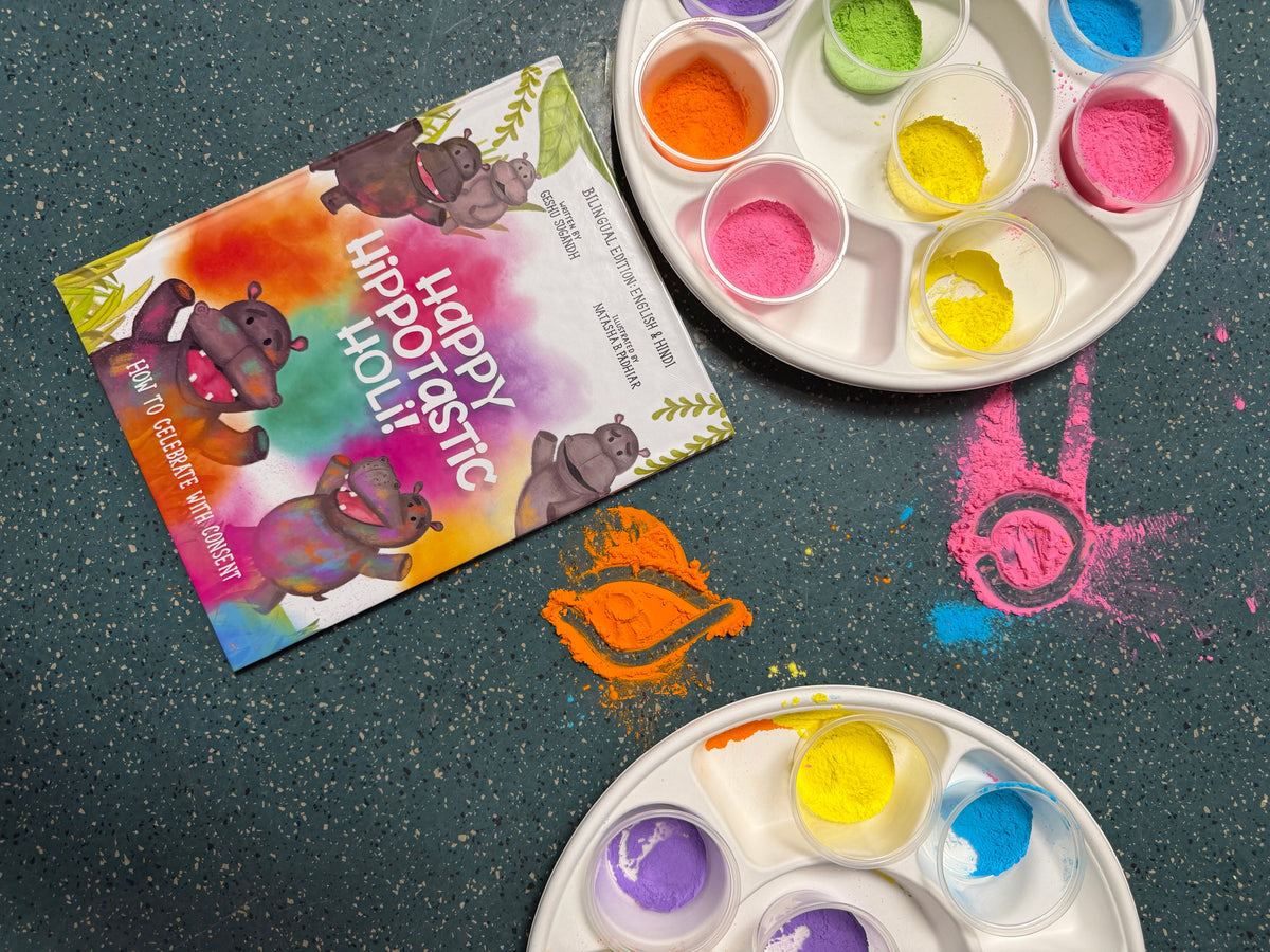 A colorful card titled 'Happy Hippoastic Holi' lies on a dark speckled surface next to two white paint palettes filled with bright powders. Orange, pink, yellow, green, purple, and blue colors are visible, with matching colored powder splatters on the surface.