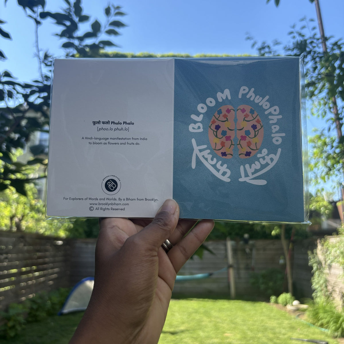 Hand holding the Brain Bloom Phulo Phalo greeting card in its clear sleeve, outdoors.