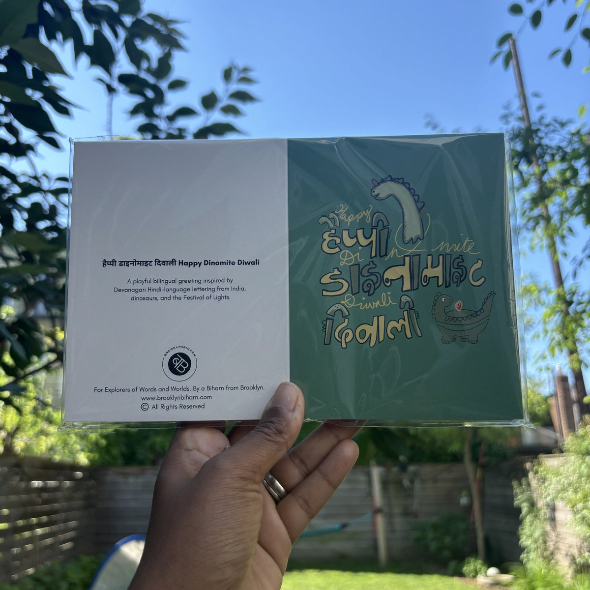 Hand holding the Happy Dinomite Diwali greeting card in its clear sleeve, outdoors.