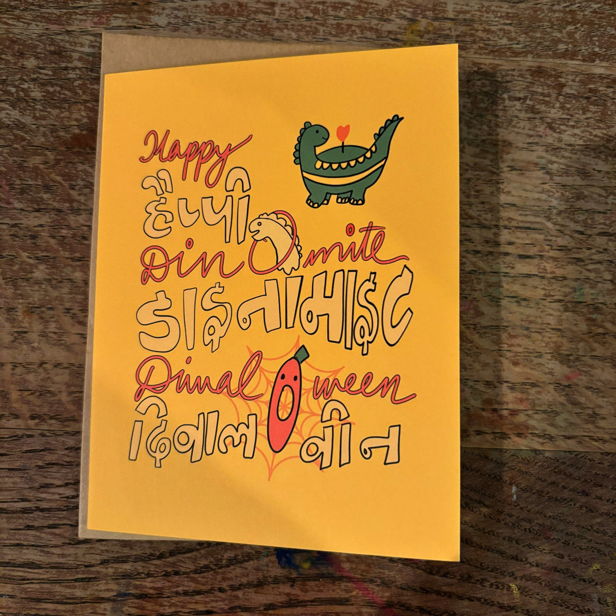 Happy Dinomite Diwal-O-ween greeting card on a wooden table with its kraft mailer.