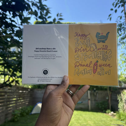 Hand holding the Happy Dinomite Diwal-O-ween greeting card in its clear sleeve, outdoors.