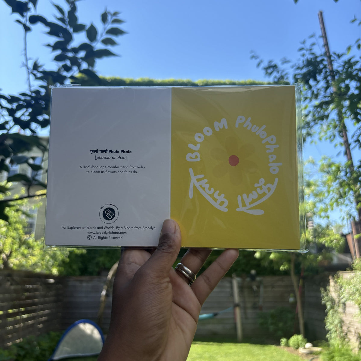 Hand holding the Flower Bloom Phulo Phalo greeting card in its clear sleeve, outdoors.