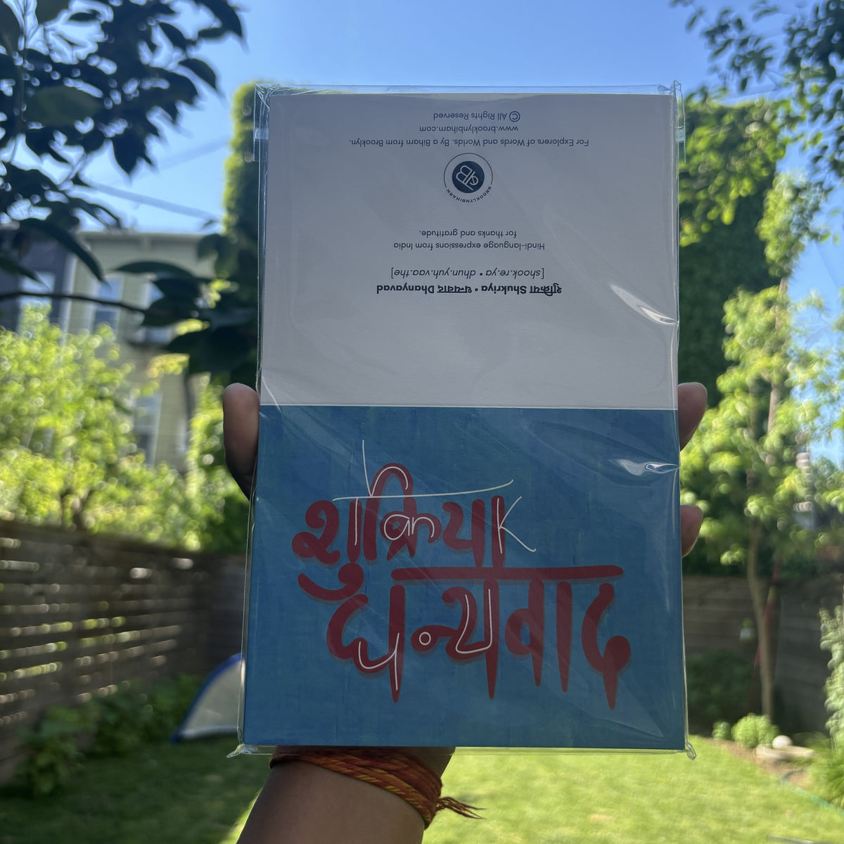 Hand holding the Shukriya Dhanyavad thank you card in its clear sleeve, outdoors.